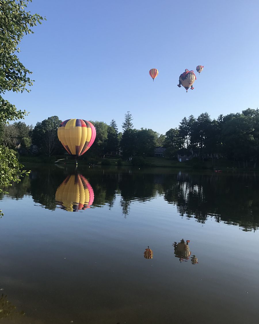 Hot-air balloons rising over a calm lake in the early morning, reflected in the water.