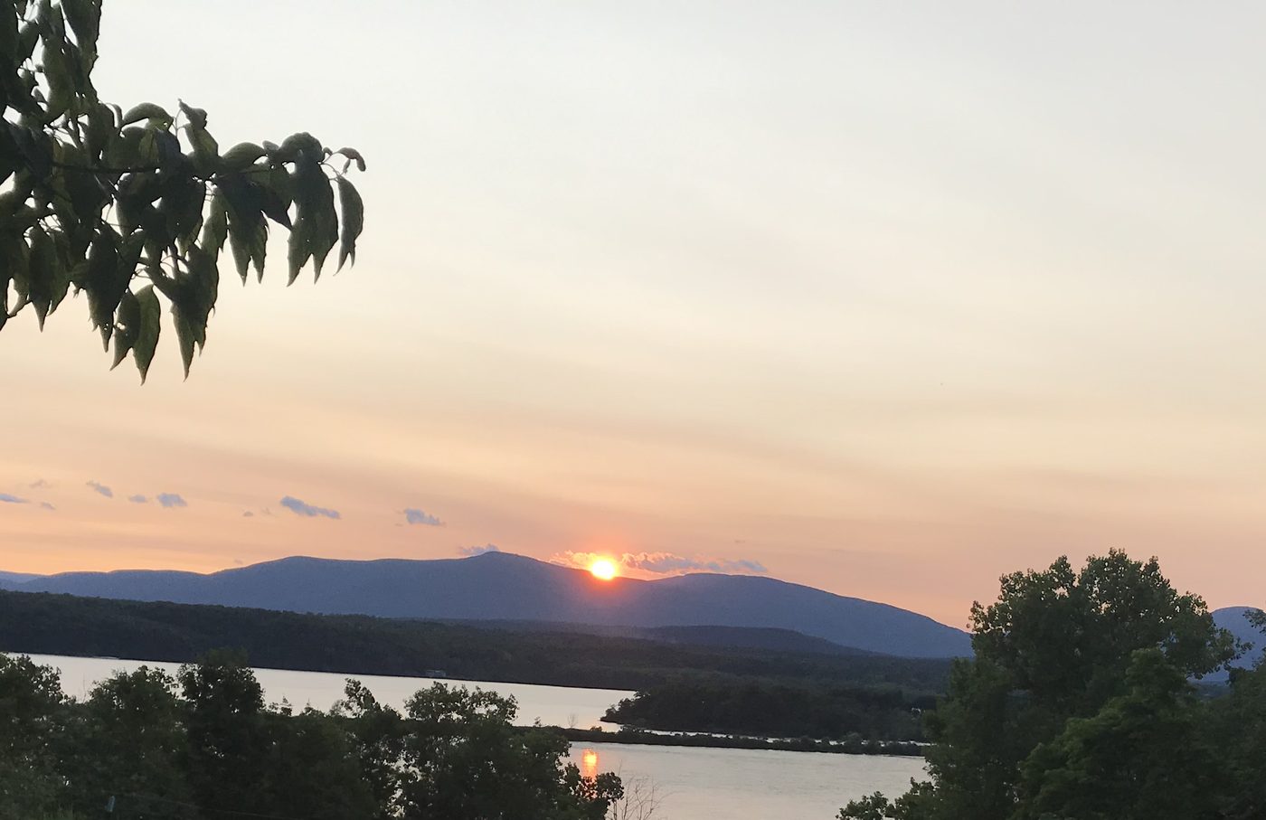 The sun setting over distant mountains beyond a wide river, framed by leaves and treetops.