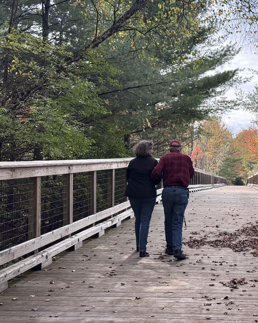 Erika and an older man walking arm in arm down a wooden boardwalk under autumn trees, seen from behind.